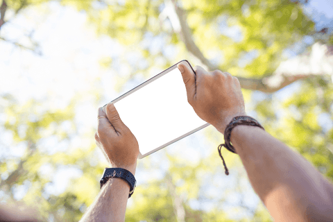 Transparent tablet held by hands amid sunlit trees