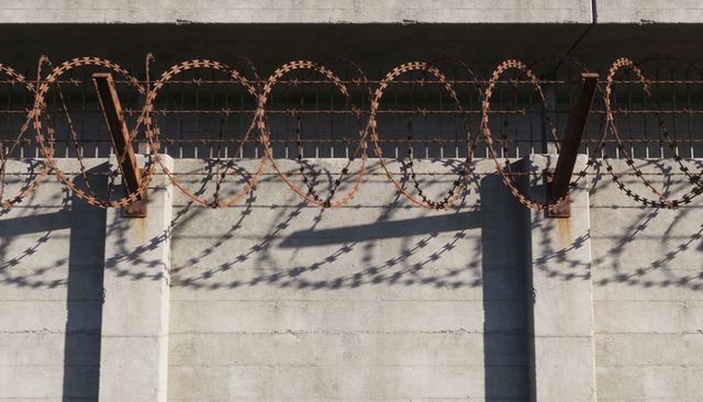Rusted concertina wire coils casting spiky shadows over weathered concrete perimeter wall