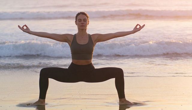 Woman practicing wide-leg squat yoga holding mudra at sunset beach with ocean waves