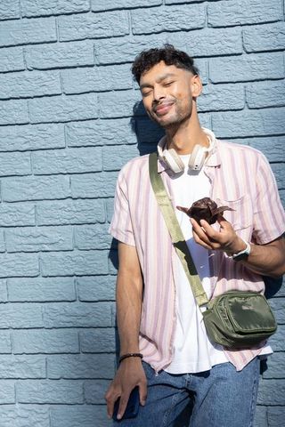 Indian man leaning on blue brick wall enjoying muffin with headphones and crossbody bag
