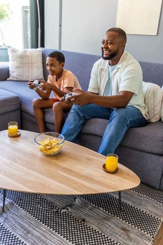 Black dad and son gaming on living room couch with snacks and juice