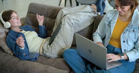 Two friends reclining on couch using laptop and tablet, casual denim jacket lifestyle