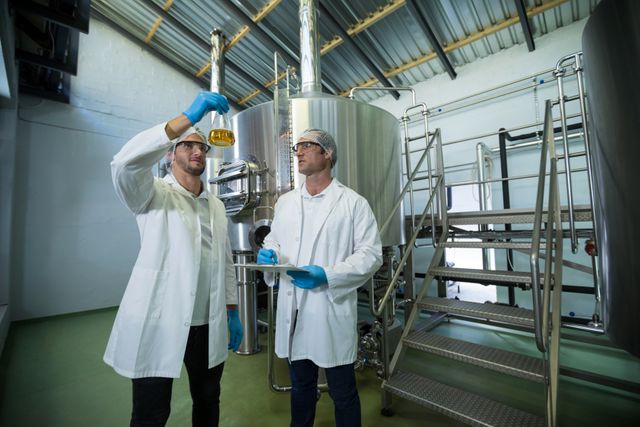 Scientists in lab coats and safety gear examining beer in a beaker at a brewery. Ideal for use in articles or advertisements related to the brewing industry, quality control, scientific research, and industrial production. Can be used to illustrate teamwork and collaboration in a professional setting.