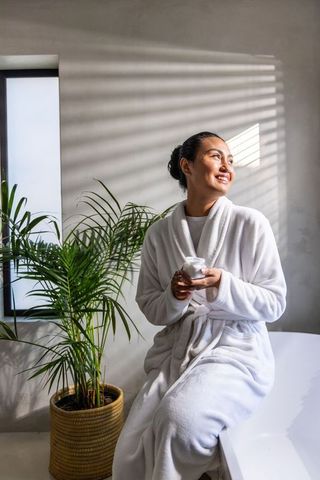 Sunlit asian woman smiling and holding moisturizer jar while sitting on bathtub edge