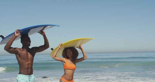 Happy biracial couple walking on beach, holding surfboard on heads and smiling. Summer, vacation, relaxation, romance, happy time.