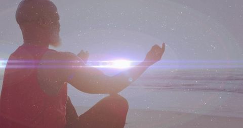 Mature man meditating on shoreline facing sunrise with dramatic lens flare and horizon
