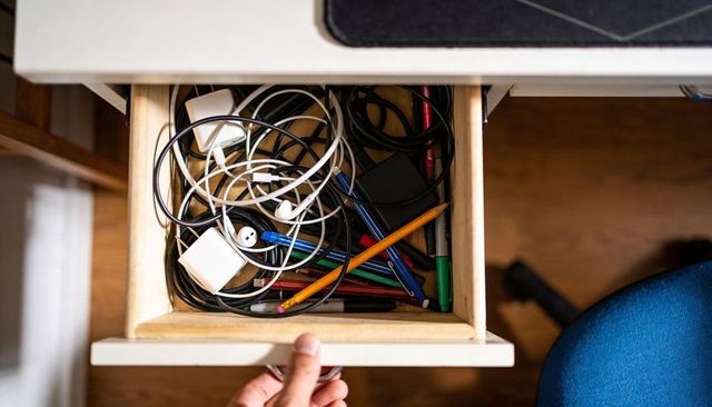 Pulling drawer revealing tangled charging cables, earbuds and colorful pens in home office