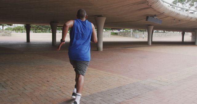 Man wearing blue tank top and gray shorts running under a concrete overpass in an urban setting. Perfect for use in fitness, sportswear, and city lifestyle promotions, as well as articles on maintaining health and fitness in urban environments.