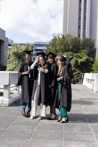 Multicultural female graduates taking selfie on campus wearing caps and gowns