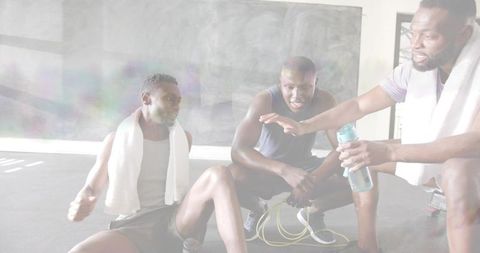 Black men resting and chatting on gym floor offering water bottle after workout