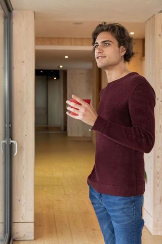 Young man standing by glass sliding door holding red cup, gazing out in wooden interior