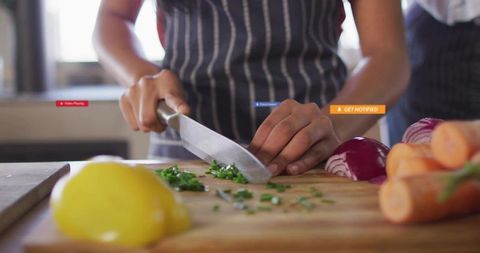 Home cook chopping fresh herbs on wooden cutting board with striped apron and vegetables