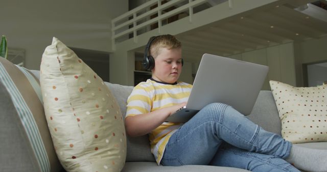 Young boy intently using laptop while wearing headphones, sitting comfortably on couch in modern home. Suitable for articles on children's use of technology, online education, remote learning, or relaxed home environments.