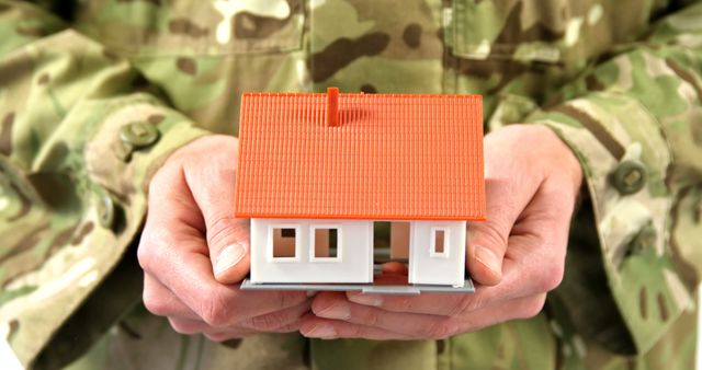 Soldier dressed in camouflage uniform holding a miniature house model in hands. Representation of home security, protection, and real estate. Suitable for use in articles or advertisements focused on housing for military personnel, property security, and veterans' aid.
