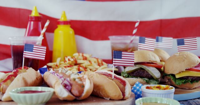 A table is laden with classic American fast food, including cheeseburgers, hot dogs, and fries, all adorned with small American flags, with copy space. The setting evokes a patriotic theme, celebrating a national holiday or American cultural event.