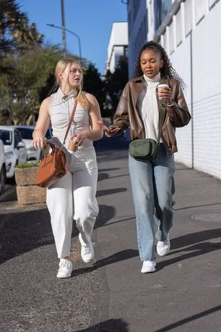 Two female friends walking and chatting on sunny urban sidewalk holding coffee cups
