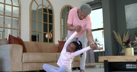 Senior african american grandmother helping granddaughter stretching in sunlit cozy living room