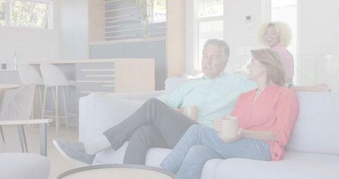Relaxing couple holding mugs on contemporary gray sofa with friend smiling in open-plan living room