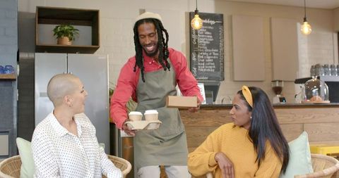 Smiling barista serving takeout coffee and pastry to two customers in cozy cafe interior