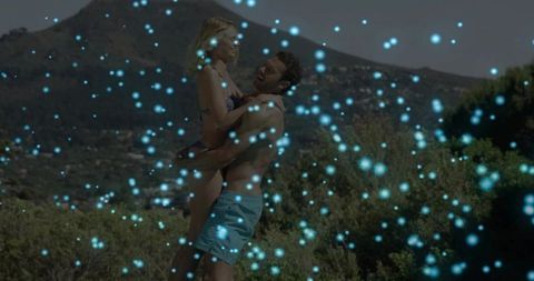 Laughing couple embracing on sunny hillside, man lifting woman, blue particle bokeh