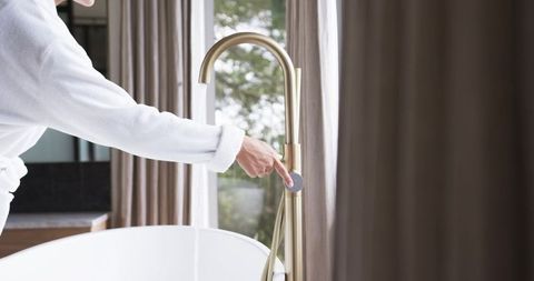 Woman in bathrobe reaching brass freestanding tub faucet with running water