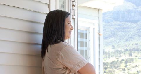Middle-aged asian woman gazing out doorway overlooking green valley, serene porch moment