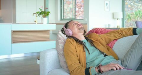 Mid adult man lounging on sofa wearing pink headphones holding smartphone in cozy living room