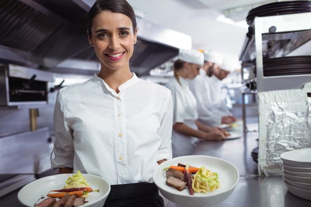 Chef holding two plates of gourmet food, smiling in a commercial kitchen. Ideal for use in articles or advertisements related to culinary arts, restaurant business, professional cooking, and team collaboration in kitchens.