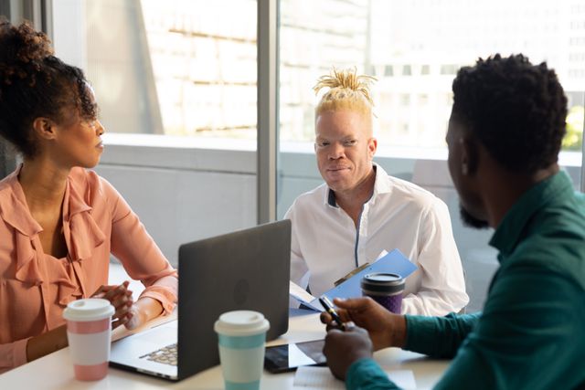 Diverse group of mid adult business professionals having a meeting in a modern office. An albino male colleague is holding documents while others listen attentively. This image is ideal for illustrating concepts of diversity, inclusion, teamwork, and corporate business environments. It can be used in articles, presentations, and marketing materials related to workplace diversity, professional collaboration, and modern office settings.