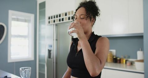 African american woman drinking water in sunlit modern kitchen wearing black cropped top