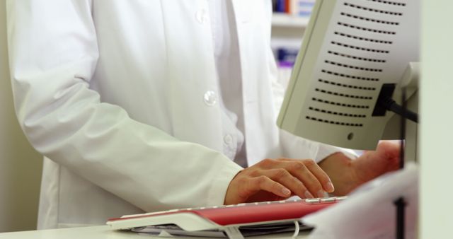 A healthcare professional in a white coat is seen typing on a computer in a medical office. This image can be used for themes related to healthcare, medical practice, administration in clinics, and digitalization in healthcare. It suits articles, websites, or promotional materials focusing on medical professionalism, healthcare services, clinic environments, and administrative tasks within healthcare settings.