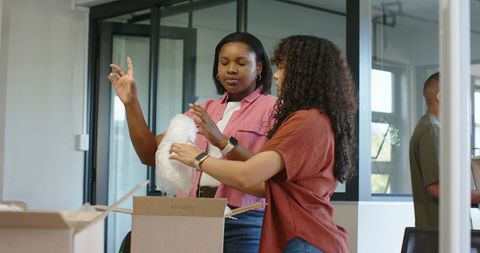 African american women packing boxes in modern office collaborating with bubble wrap