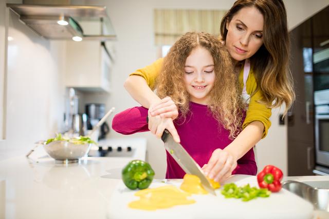 Mother assisting daughter in cutting vegetables in a modern kitchen. Ideal for illustrating family bonding, healthy eating habits, parenting, and home cooking. Perfect for use in articles, blogs, and advertisements related to family life, nutrition, and culinary education.