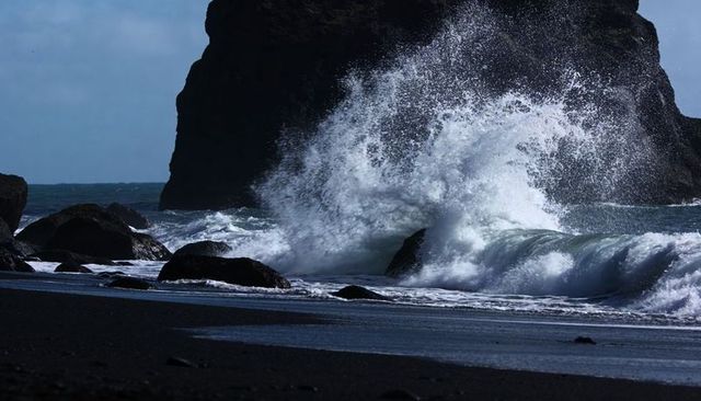 Dark Volcanic Beach with Crashing Wave and Towering Spray against Sea Stack