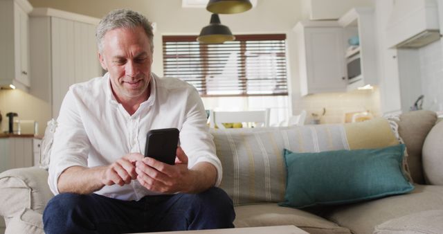 Happy caucasian man sitting on sofa in living room, using smartphone. Spending time at home alone.