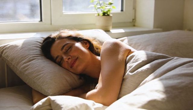 Woman sleeping peacefully on sunlit bed by window with striped pillow and green plant