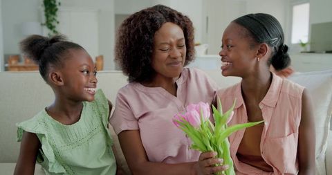 Mother receiving pink tulip bouquet from smiling daughters on couch, family togetherness