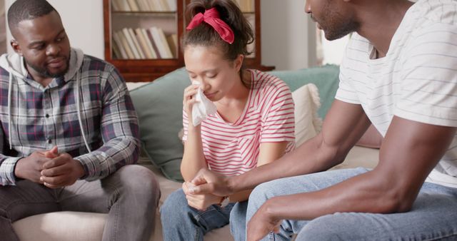 Diverse group of friends comforting grieving woman using tissues while sitting on a couch at home. Useful for topics related to mental health, emotional support, friendship, or grieving process. Can be used in articles, blogs, and advertisements about support systems, counseling, and personal relationships.