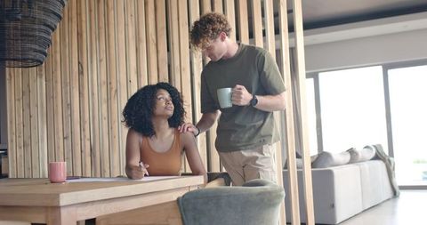 Sunlit modern living moment man offering coffee as partner pauses writing at wooden table