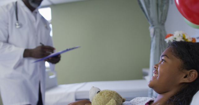 Doctor writing on clipboard while smiling young girl holds teddy bear in hospital bed. Ideal for healthcare, pediatric care, children in hospital settings, medical services, emotional comfort during treatment visuals.