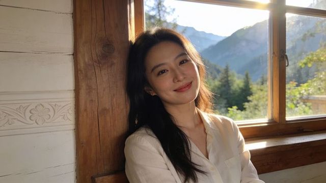 Sunlit asian woman leaning against wooden beam in rustic mountain cabin smiling peacefully