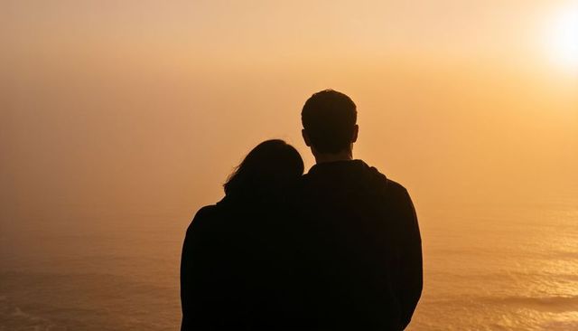 Silhouetted couple watching golden ocean sunset leaning together on coastal cliff