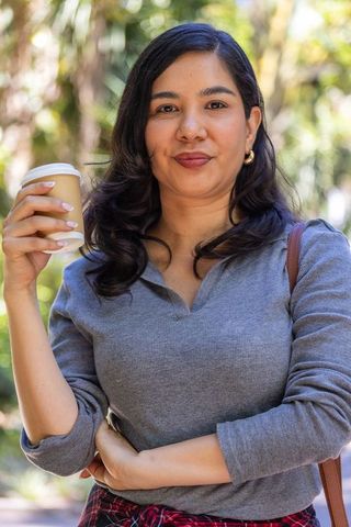 Young woman holding takeaway coffee cup in sunlit park, casual sweater and tied plaid shirt