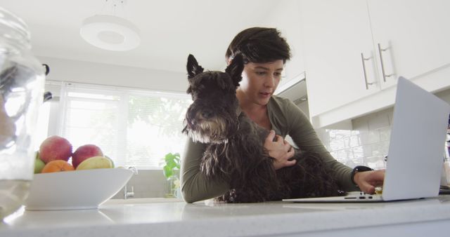 Scene emphasizes work-life balance with a woman working on a laptop while holding a dog in a bright kitchen. Perfect for illustrating remote work, pet care, kitchen activities, or balancing household duties and job responsibilities.
