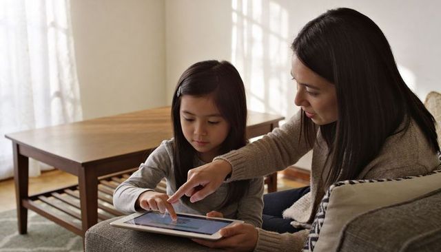 Mother teaching daughter using tablet on sofa in sunlit living room for digital learning