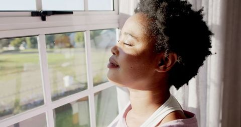 African American Woman Enjoying Sunlight by Window, Peaceful Reflection at Home