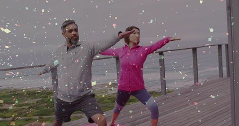 Couple practicing warrior yoga pose on coastal boardwalk with confetti effect
