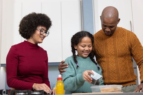 Diverse family preparing breakfast together, child pouring ingredients and smiling