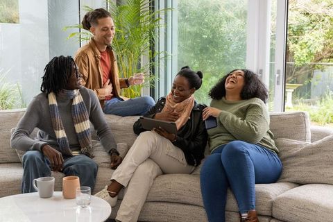Multicultural friends laughing and sharing tablet on cozy sectional sofa at home