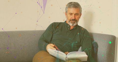 Senior man reading book on gray sofa with digital network overlay, relaxed reader